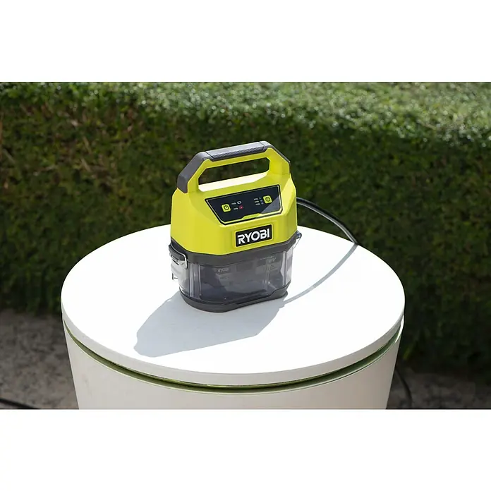 Ryobi ONE+ Schmutzwasserpumpe - Max. Fördermenge: 4.200 l/h - Nahaufnahme eines tragbaren Ryobi-Akkuladegeräts auf einem runden, weißen Tisch im Freien, mit üppigem Grün im Hintergrund.