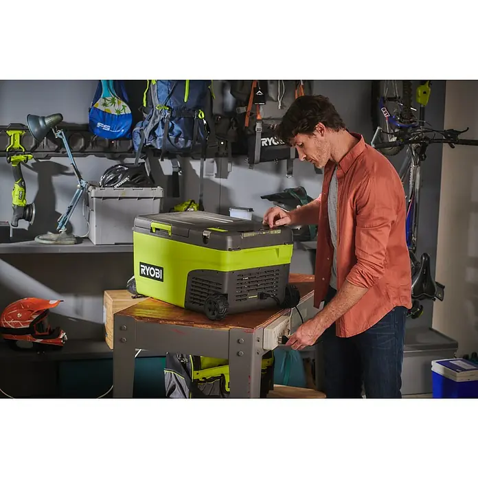 Ryobi ONE+ Akku-Kühlbox Hybrid RY18CB23A-0 - L x B x H: 72,5 x 42 x 49,5 cm, Fassungsvermögen: 23 l - Ein RYOBI Kühlbox steht auf einer Werkbank, während ein Mann sie an eine Steckdose anschließt. Die Kühlbox ist grün und schwarz.