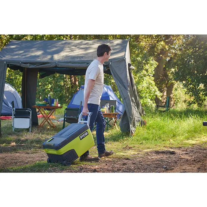 Ryobi ONE+ Akku-Kühlbox Hybrid RY18CB23A-0 - L x B x H: 72,5 x 42 x 49,5 cm, Fassungsvermögen: 23 l - Ein Mann transportiert eine gelbe Ryobi Kühlbox auf Rädern auf einem Campingplatz.