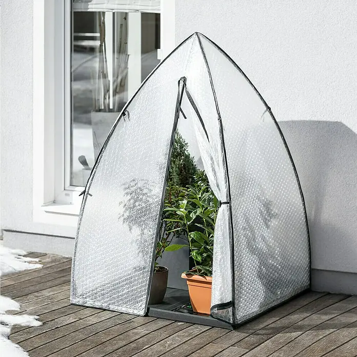 Windhager Winterschutzzelt Yukon - Luftpolsterfolie, L x B x H: 1,2 x 1,2 x 1,8 m - Ein kleines Gewächshaus mit Pflanzen auf einer Holzterrasse, vor einer weißen Hauswand und einem Fenster.