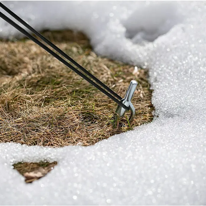 Windhager Winterschutzzelt Yukon - Luftpolsterfolie, L x B x H: 1,2 x 1,2 x 1,8 m - Nahaufnahme eines silbernen Zeltnagels, der mit einer schwarzen Schnur im grasbewachsenen Boden befestigt ist, teilweise mit Schnee bedeckt.