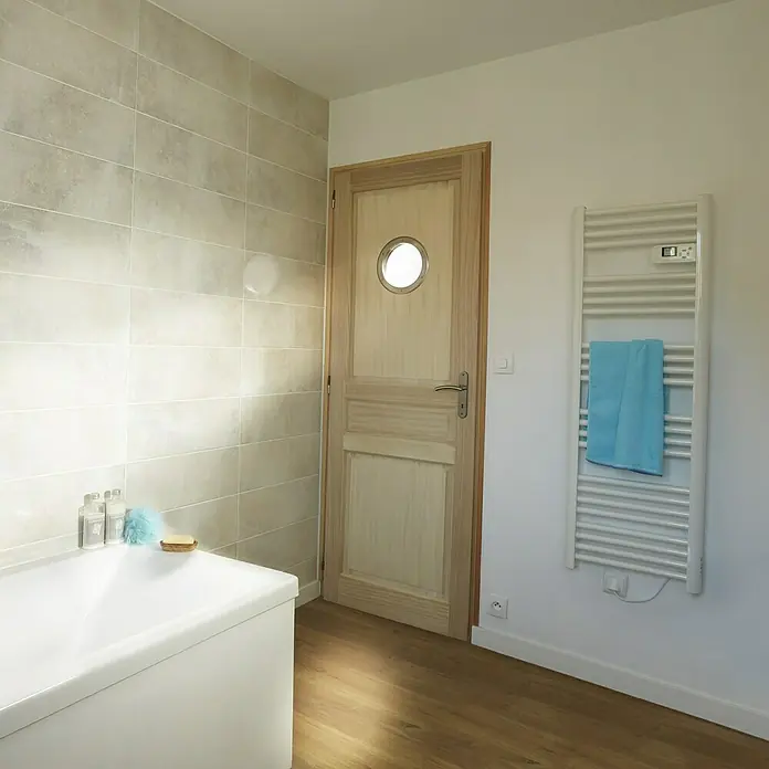 Mottez Bullaugen-Glas - Glas, Milchglas, Passend für: Mottez Bullaugen Ø 26 cm - Ein Badezimmer mit einer Badewanne, einem Handtuchwärmer mit blauem Handtuch und einer Holztür mit Bullauge.