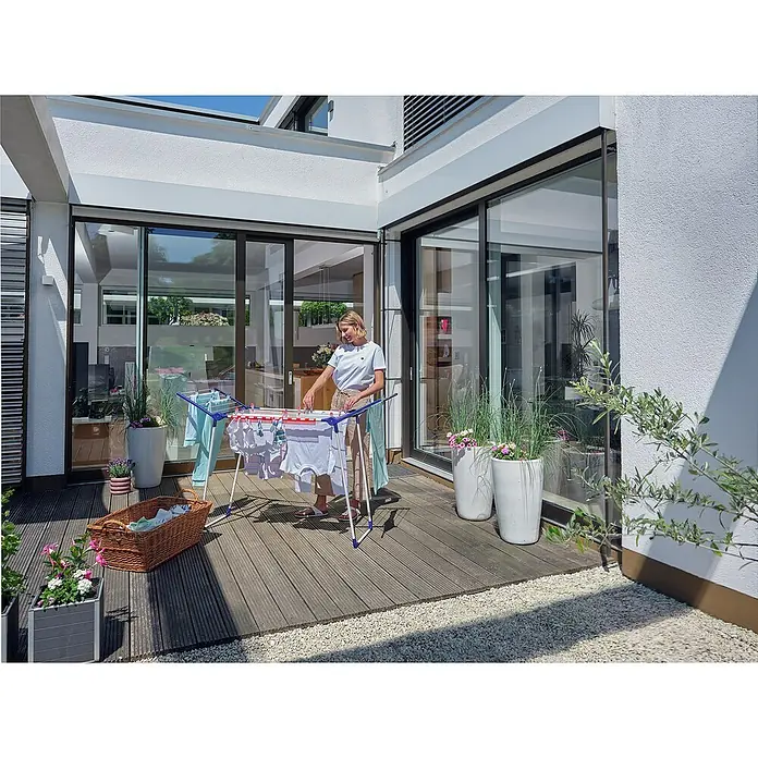 Leifheit Flügel-Wäscheständer Pegasus 150 Solid Slim - Länge Wäscheleine: 15 m, L x B x H: 87 x 55 x 157 cm, Weiß/Blau - Eine Frau steht auf einer Terrasse und hängt Wäsche auf einen Wäscheständer. Zu sehen sind auch Blumen und Terrassendielen.