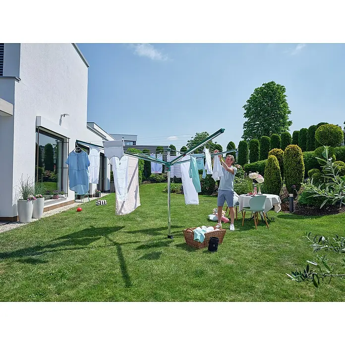 Leifheit Wäschespinne Linomatic 600 Deluxe Cover - Länge Wäscheleine: 60 m, Geeignet für Anzahl Waschmaschinenfüllungen: 6 Stk. - Wäschespinne mit Wäsche im Garten, ein Mann hängt die Wäsche auf. Im Hintergrund ein Haus und Bäume.