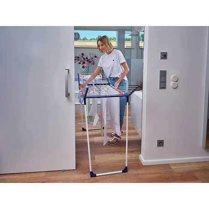 Leifheit Flügel-Wäscheständer Pegasus 150 Solid Slim - Länge Wäscheleine: 15 m, L x B x H: 87 x 55 x 157 cm, Weiß/Blau - Frau beim Wäscheaufhängen auf einem blauen und weißen Wäscheständer im Innenbereich.
