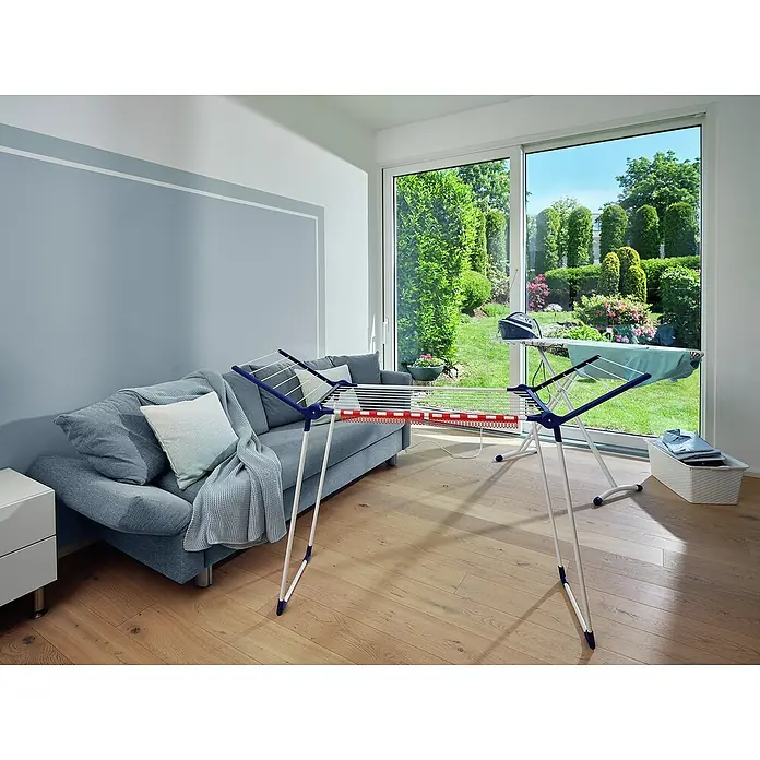 Leifheit Flügel-Wäscheständer Pegasus 150 Solid Slim - Länge Wäscheleine: 15 m, L x B x H: 87 x 55 x 157 cm, Weiß/Blau - Wäscheständer im modernen Wohnraum. Der robuste Ständer mit roten Akzenten steht vor einem Sofa und bietet viel Platz zum Trocknen von Kleidung.
