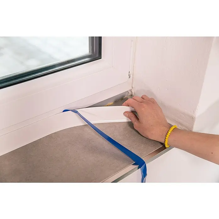 Schellenberg Flachleiste - 50 m x 30 mm x 1,5 mm, Weiß - Nahaufnahme einer Hand, die eine Fensterbank montiert, mit blauem Klebeband zur Befestigung. Das neue Fensterbrett ist aus weißem Material gefertigt.