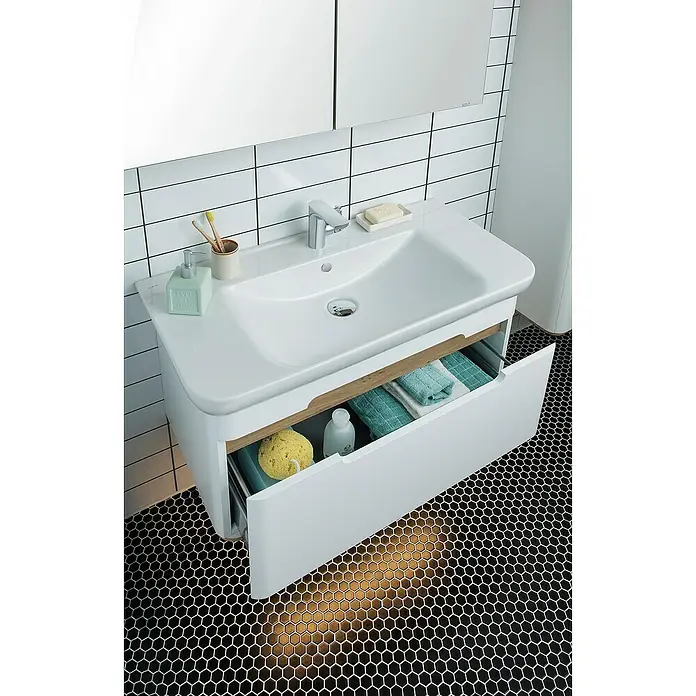 VitrA Sento Badmöbel-Set - 98 cm, Weiß, Griffmulde - Elegantes Waschbecken mit offenem Unterschrank. Blick auf Utensilien wie Handtücher, Schwamm und Lotion. Schwarze Sechseckfliesen am Boden.