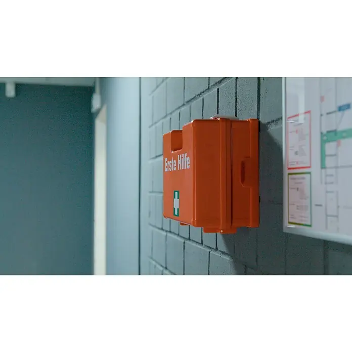 Leina-Werke Erste-Hilfe-Koffer Quick - DIN 13157, Mit Wandhalterung, Orange - Nahaufnahme einer orangefarbenen Erste-Hilfe-Box an einer Wand, die mit einem Erste-Hilfe-Logo und Text versehen ist.