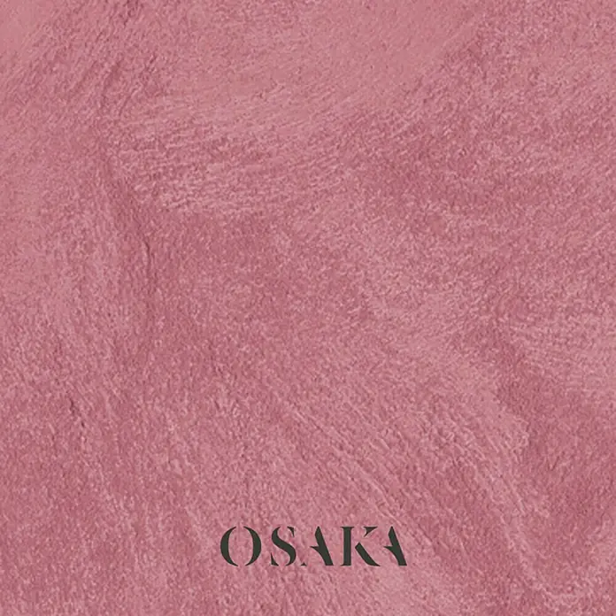 Osaka Pintura para efectos decorativos Arena - 5 l, Rojo, Mate - Un primer plano de una pared rosa con la palabra "OSAKA" en letras oscuras. La atención se centra en la textura de la pared y el diseño sencillo.