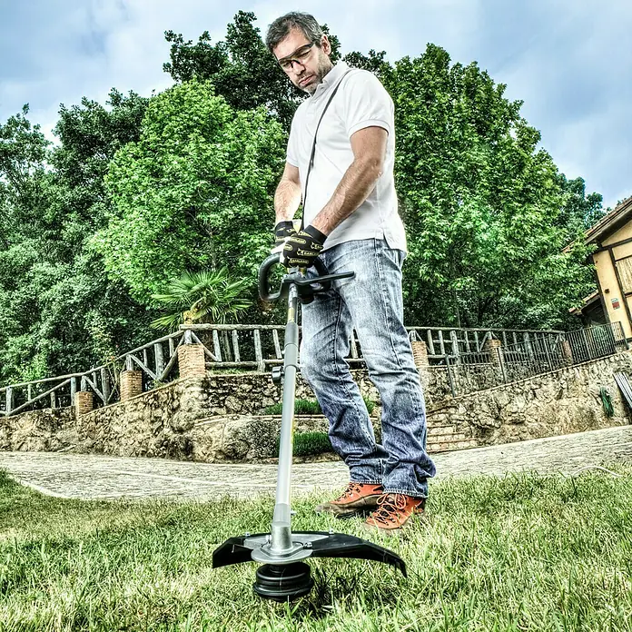 Garland Desbrozadora de disco eléctrica Best 100 - 1.000 W, Ancho de corte hilo: 38 cm, Ancho de corte cuchilla: 23 cm - Un hombre está usando una desbrozadora para cortar el césped. La desbrozadora es plateada y negra y tiene un mango para sujetarla.