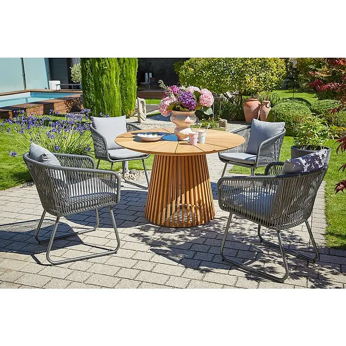 Sunfun Gartenmöbel-Set Osby/Maja - 5 -tlg., Teak/Aluminium/Stahl/Kunststofffaser-Strings, Teak/Grau - Ein runder Gartentisch aus Holz mit vier Stühlen mit grauen Kissen steht auf einer Terrasse mit viel Grün im Hintergrund.