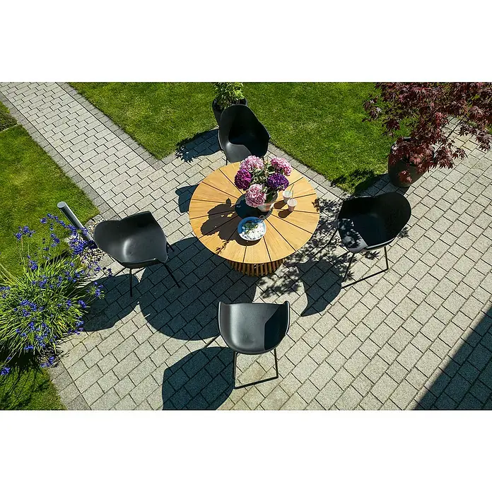 Sunfun Gartenmöbel-Set Osby/My - 5 -tlg., Teak/Aluminium/Kunststoff, Teak/Schwarz - Gartenmöbel-Set mit einem runden Holztisch und vier schwarzen Stühlen auf einer gepflasterten Terrasse mit Blumen und Bäumen.