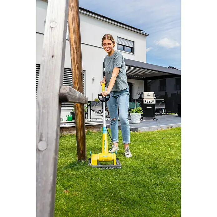 Gloria MultiBrush Grasscherenaufsatz FineCut - Passend für: MultiBrush PLUS Geräte - Eine Frau benutzt eine gelbe MultiBrush Bürste, um Rasen zu reinigen. Das Gerät ist auf Rasen platziert, im Hintergrund ein Haus.