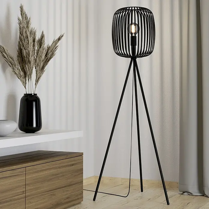 Eglo Romazzina Lámpara de pie - 40 W, Altura: 140,5 cm, Negro, E27 - Una elegante lámpara de pie trípode negra con una pantalla en forma de jaula y una bombilla. Elementos decorativos al fondo.