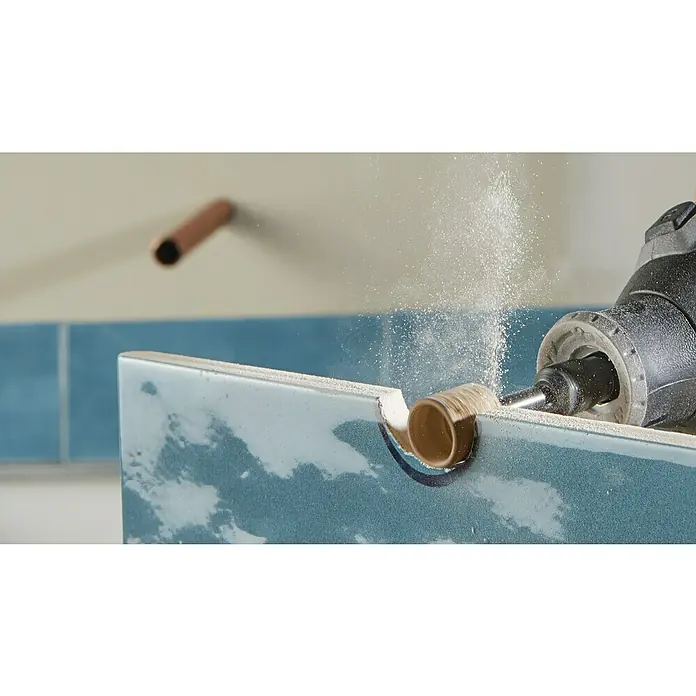 Dremel Schleifband / Lochbohrer 408DM (2-in-1)Körnung: 60 - Ein Dremel-Werkzeug schneidet ein Loch in eine blaue Keramikfliese, wobei Staub aufgewirbelt wird. Eine weitere Fliese mit Rohr ragt aus der Wand.