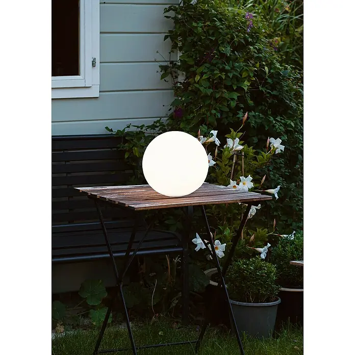 Trio Lighting Lámpara de diseño para exterior Bahamas - Diámetro: 25 cm, IP44 - Escena de jardín con una lámpara globo redonda y luminosa en una mesa de madera, rodeada de plantas y un edificio al fondo.