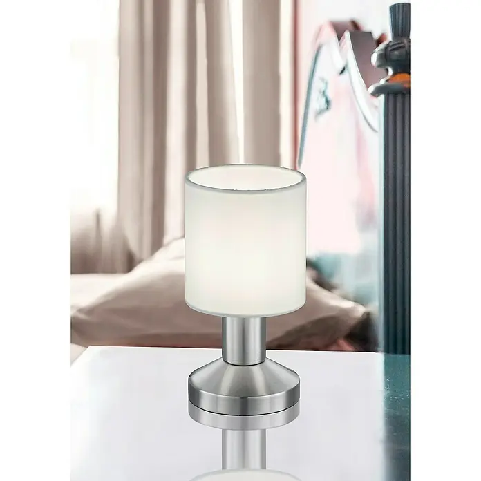 Trio Lighting Lámpara de sobremesa Garda - 25 W, L x An x Al: 9,5 x 9,5 x 18 cm, Blanco, E14 - Primer plano de una moderna lámpara de noche con pantalla cilíndrica y base plateada, que emite una luz suave en el dormitorio.
