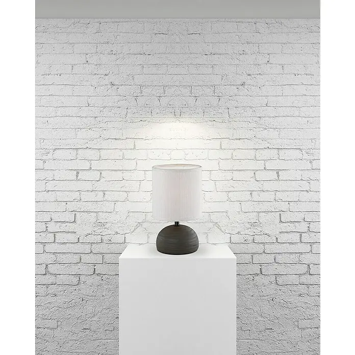 Trio Lighting Lámpara de sobremesa Luci - 40 W, Ø x Al: 14 x 23 cm, Negro, E14 - Una lámpara de mesa elegante con base gris y pantalla blanca sobre un pedestal blanco frente a una pared de ladrillos.