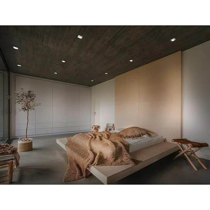 Trio Lighting Set de focos empotrables Jura - Blanco, Diámetro: 8 cm, Cuadrado - El dormitorio minimalista cuenta con una cama de plataforma baja con una manta marrón clara, detalles de madera e iluminación empotrada en el techo.