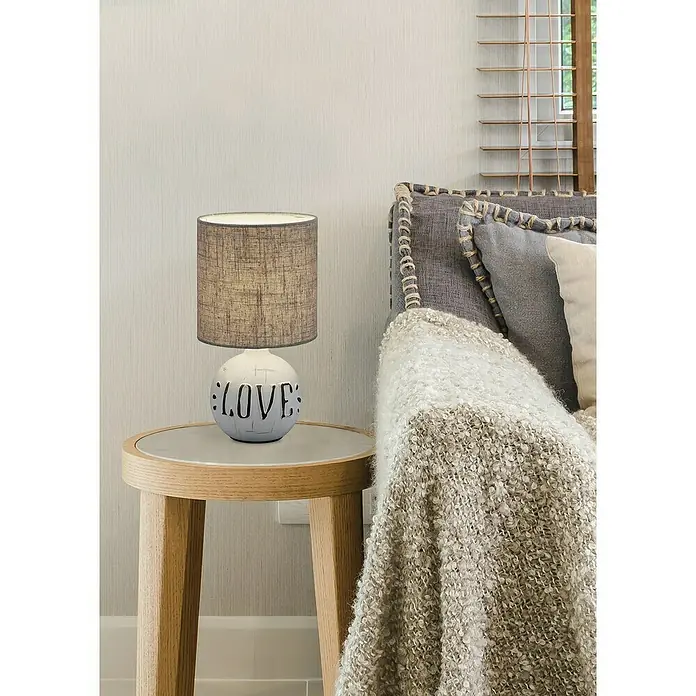 Reality Leuchten Lámpara de sobremesa redonda Esna Love - Love, 40 W, Ø x Al: 165 mm x 31 cm, Gris/Marrón, E14 - Primer plano de una lámpara de noche con un diseño de 'Love' sobre una mesa redonda de madera junto a una cama acogedora.