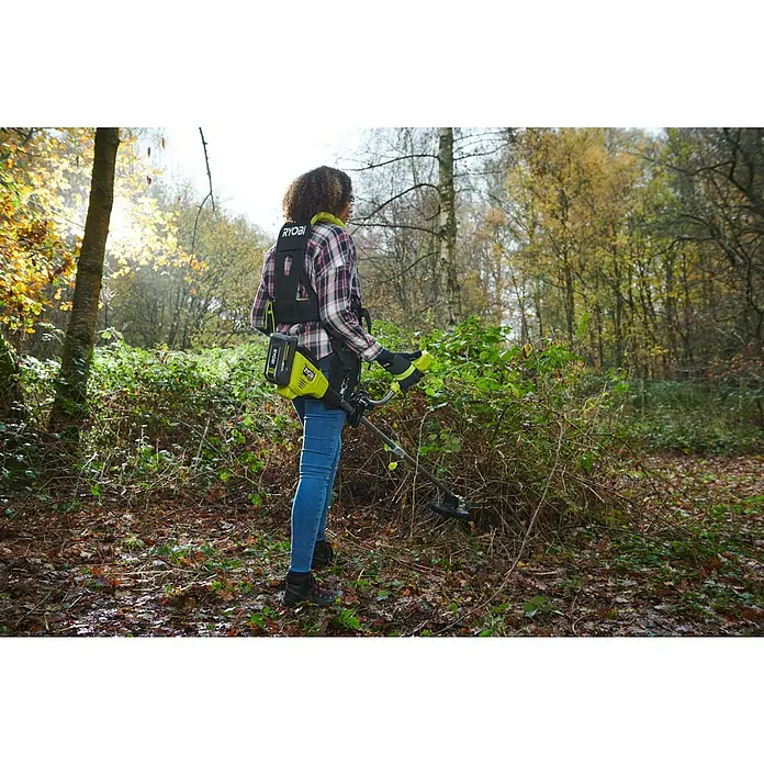 Ryobi Max Power Akku-Freischneider RY36BCX30A-9 - 36 V, Ohne Akku, Schnittbreite Faden: 40 cm, Schnittbreite Messer: 30 cm - Eine Frau benutzt einen Ryobi Freischneider mit Akku in einem Wald, um Gestrüpp zu entfernen. Der Freischneider ist mit einem Tragegurt ausgestattet.