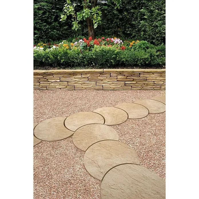 SAS Trittstein Luna - 34 x 47 x 3 cm, Arena, Beton - Ein Gartenweg mit beigen, halbkreisförmigen Trittsteinen, eingebettet in roten Kies vor einer Steinmauer und üppiger Bepflanzung.