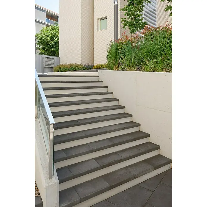 SAS Tessera Poolrandplatte - 32,8 x 50 x 6 cm, Anthrazit, Beton - Moderne Außentreppe mit grauen Stufen und weißem Geländer vor einem Gebäude. Die Treppe wird von grünen Pflanzen gesäumt.