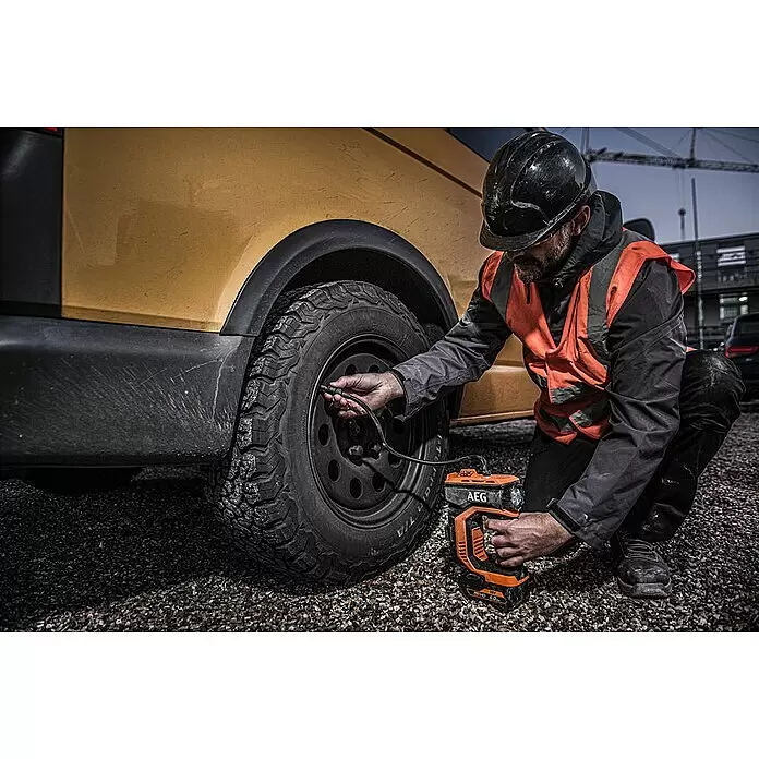 AEG Pro 18V Compresor de batería BK 18C - 18 V, Sin batería, 10,3 bar - Un hombre infla un neumático sucio de un vehículo con un inflador de neumáticos a batería AEG naranja y negro sobre grava.