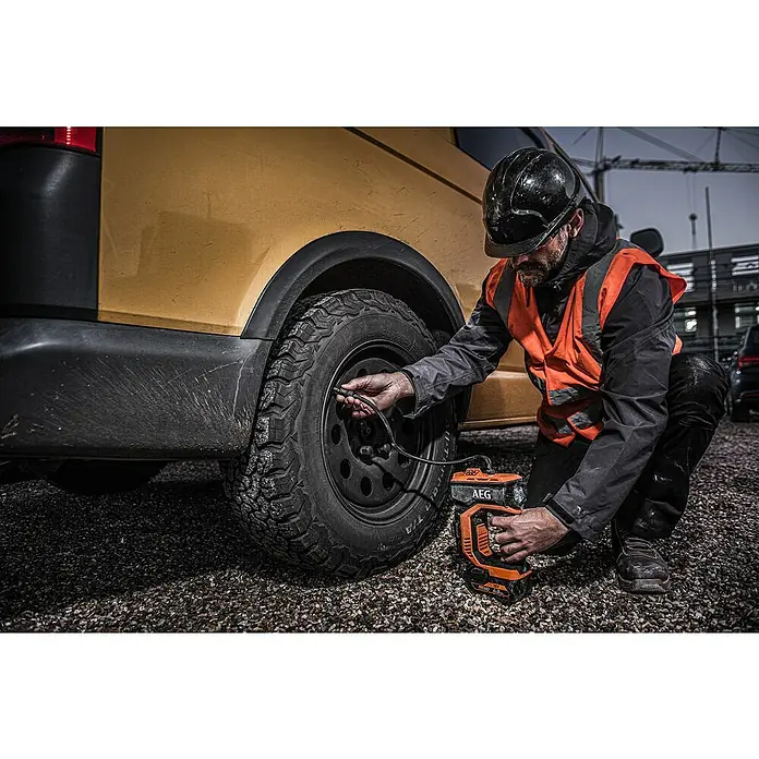 AEG Pro 18V Akku-Kompressor BK 18C - 18 V, Ohne Akku, 10,3 bar - Ein Mann befüllt einen Reifen mit einem AEG Luftkompressor. Das orangefarbene Gerät steht auf einem Kiesweg neben einem gelben Van.