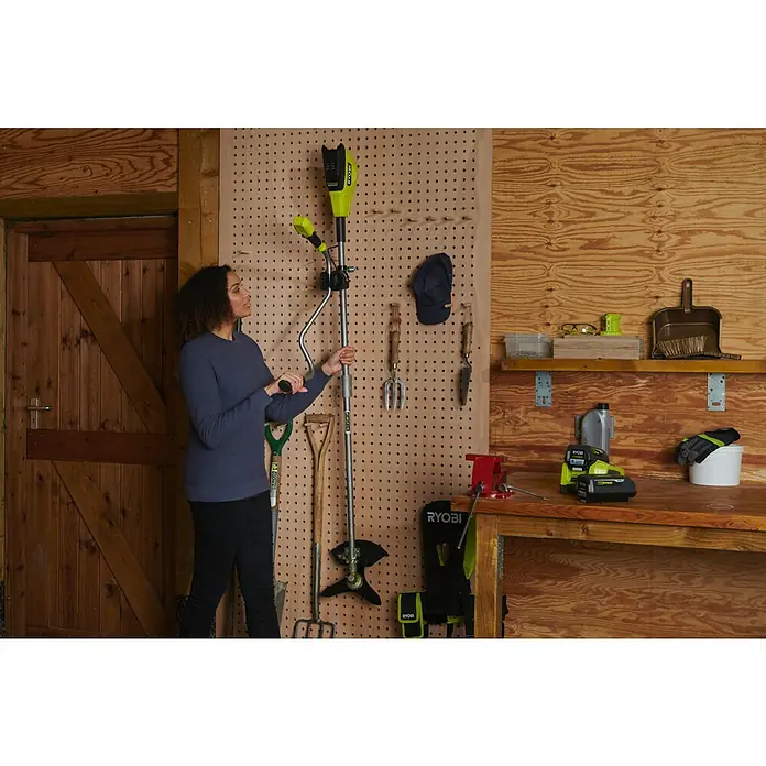 Ryobi Max Power Akku-Freischneider RY36BCX30A-9 - 36 V, Ohne Akku, Schnittbreite Faden: 40 cm, Schnittbreite Messer: 30 cm - Eine Frau betrachtet einen RYOBI Akku-Rasentrimmer, der an einer Lochwand in einer Werkstatt hängt. Das Werkzeug ist ein praktischer Helfer.