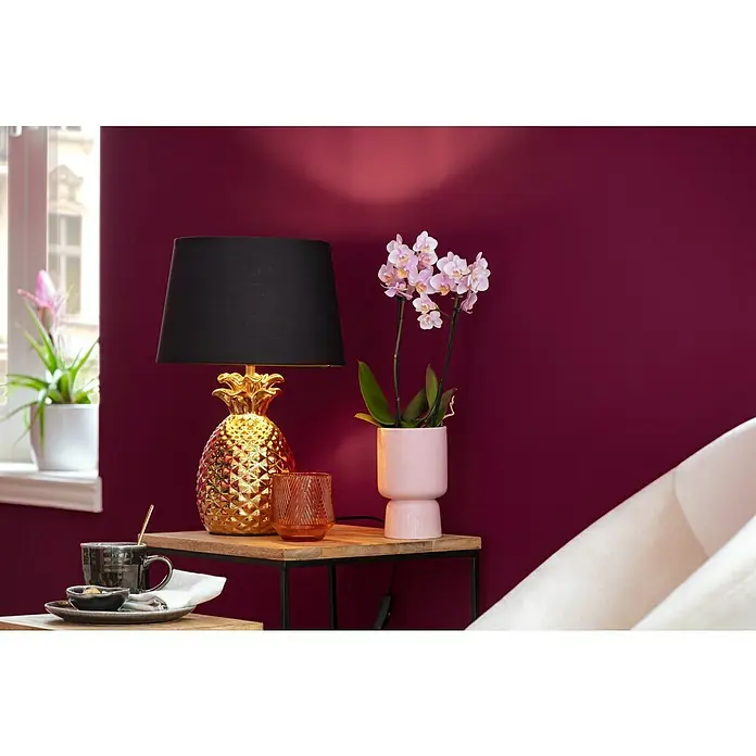 Reality Leuchten Tischleuchte Pineapple - 60 W, E27 - Nahaufnahme einer goldenen Ananas-Tischlampe mit schwarzem Lampenschirm, einer Orchidee und einer Tasse auf einem Holztisch vor einer burgunderroten Wand.