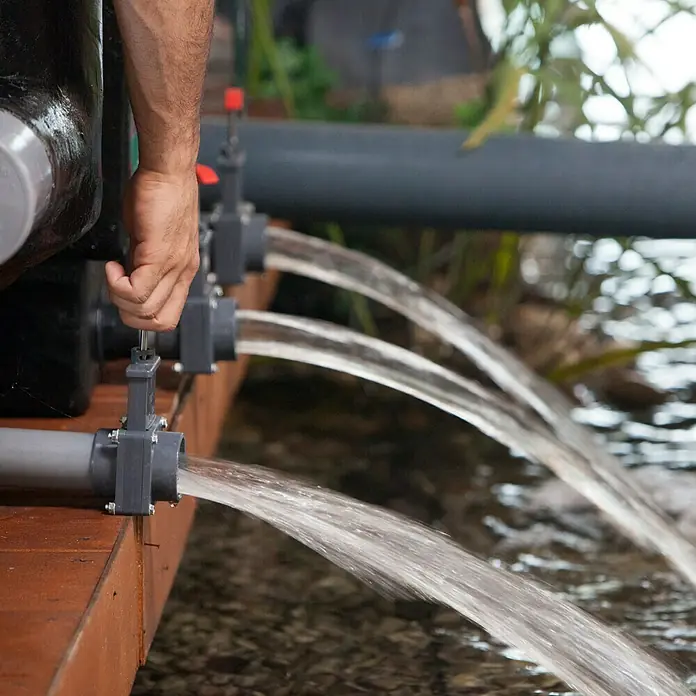 Heissner Durchlauffilter Apollo XL Pro F322 - Teichgröße: 80 m³ - Wasserauslass: Eine Hand öffnet ein Ventil, aus dem Wasser in drei Strömen austritt, Teil einer Aquakulturanlage im Freien.