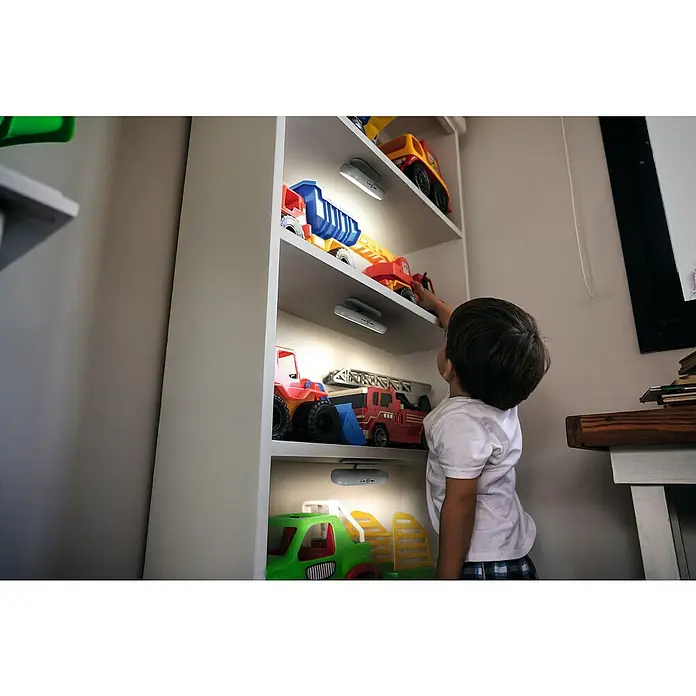 Ledvance LED-Unterbauleuchte Linear Mobile Tilt Sensor - Länge: 18,6 cm, Kaltweiß - Kind erreicht nach Spielzeugtrucks in einem weißen Spielzeugregal mit eingebauten Leuchten.\n