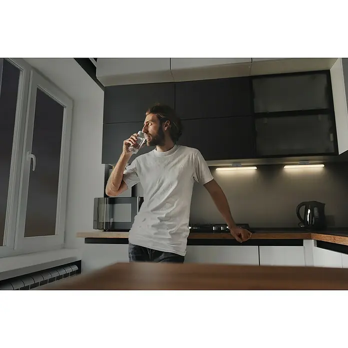 Ledvance LED-Unterbauleuchte Linear Swivel - Länge: 35 cm, Warmweiß - Ein Mann in einem weißen T-Shirt steht in einer Küche und trinkt ein Glas Wasser, mit modernen Schränken und Geräten im Hintergrund.