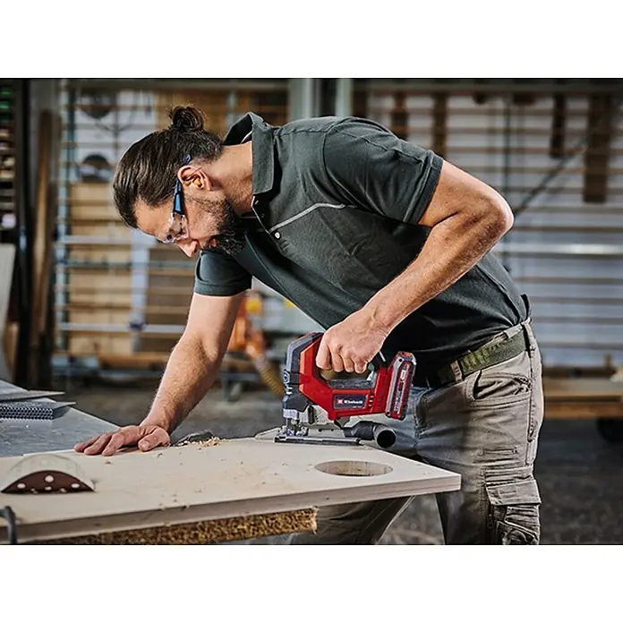 Einhell Professional PXC 18V Sierra de calar a batería con acción pendular - 18 V, Número de baterías: Sin batería, 3.500 /min - Un hombre usa una sierra de calar inalámbrica Einhell para cortar un agujero en una tabla de madera, mientras usa gafas de seguridad y protección auditiva.