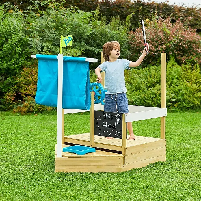 TP Toys Sandkasten Ahoy - 180 x 92 x 118 cm, Holz, Natur/Weiß, Blau - Ein Kind spielt auf einem hölzernen Spielschiff mit blauem Segel und Steuerrad im Garten. Das Schiff hat eine Tafel mit der Aufschrift "Ship Ahoy!".