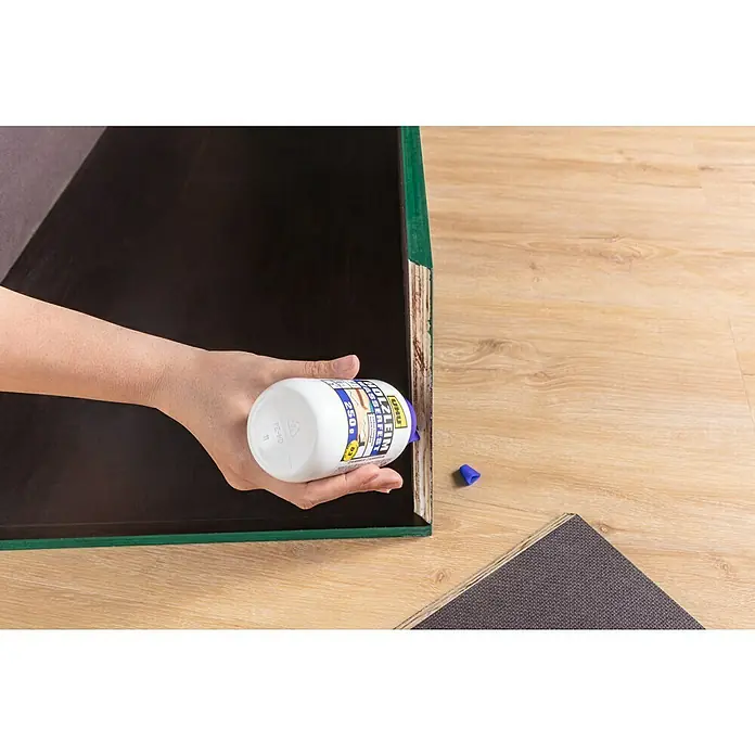 UHU Holzleim Wasserfest - 750 g, Lösemittelfrei - Nahaufnahme einer Hand, die Holzleim auf ein Holzprojekt aufträgt, wobei die Flasche des Holzleims klar im Fokus steht.