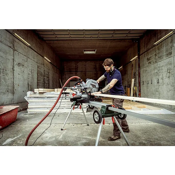 Metabo Kapp- & Gehrungssäge KGS 305 M - 1.600 W, Durchmesser Sägeblatt: 305 mm, Leerlaufdrehzahl: 3.700 U/min - Ein Mann bedient eine Metabo Kapp- und Gehrungssäge KGS 216 M auf einem Montageständer, um Holz zu schneiden.