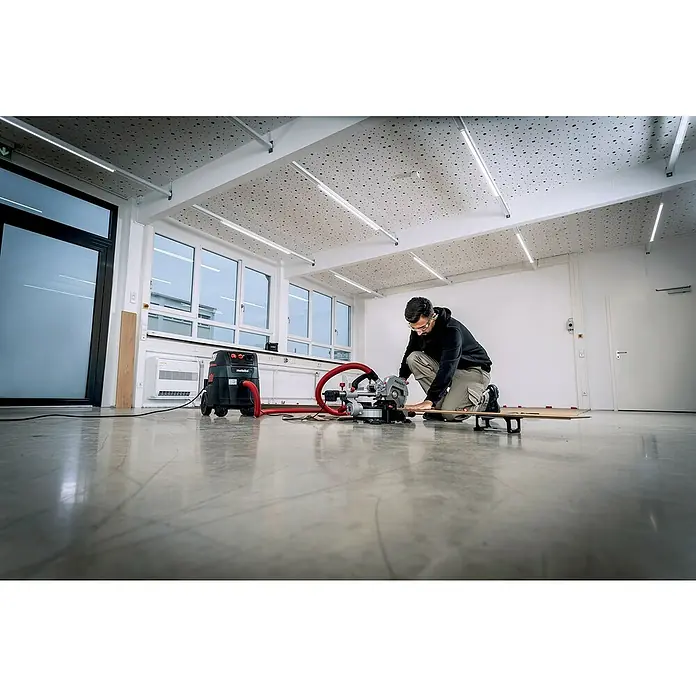 Metabo Kapp- & Gehrungssäge KGS 216 M Set - 1.500 W, Durchmesser Sägeblatt: 216 mm, Leerlaufdrehzahl: 5.000 U/min - Ein Handwerker kniet vor einer Metabo Kappsäge, die an einen Metabo Staubsauger angeschlossen ist, in einem hellen Raum.