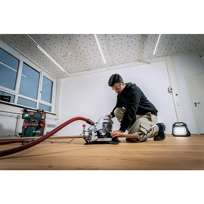 Metabo Kapp- & Gehrungssäge KS 216 M - 1.100 W, Durchmesser Sägeblatt: 216 mm, Leerlaufdrehzahl: 5.000 U/min - Ein Mann kniet vor einer Metabo Kappsäge mit Staubabsaugung, um ein Holzbrett zu schneiden, Schutzbrille tragend.