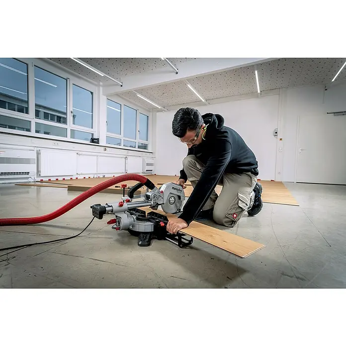 Metabo Kapp- & Gehrungssäge KGS 216 M Set - 1.500 W, Durchmesser Sägeblatt: 216 mm, Leerlaufdrehzahl: 5.000 U/min - Ein Mann kniet und schneidet ein Holzbrett mit einer Metabo Kappsäge in einem hellen Raum. Schutzkleidung wird getragen und ein Absaugschlauch ist angeschlossen.