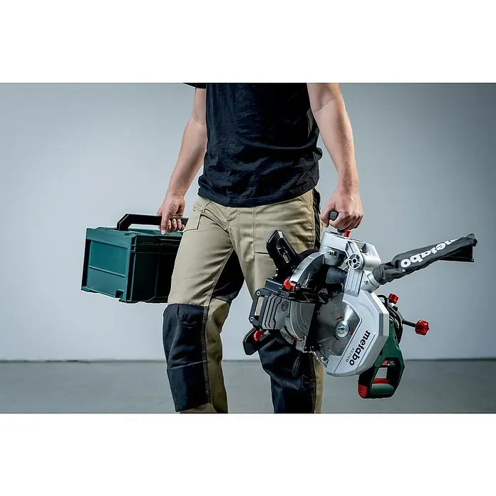 Metabo Kapp- & Gehrungssäge KS 216 M - 1.100 W, Durchmesser Sägeblatt: 216 mm, Leerlaufdrehzahl: 5.000 U/min - Eine Person trägt eine Metabo Kappsäge KS 216 M und einen Werkzeugkoffer. Ideal für präzise Schnitte und den mobilen Einsatz auf Baustellen.