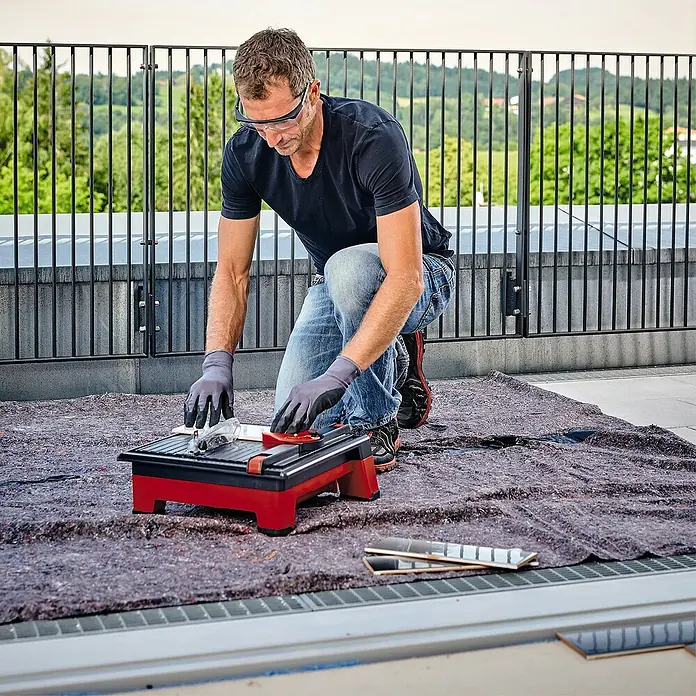 Einhell Power X-Change 18V Akku-Fliesenschneider TE-TC 18/11518 V, Ohne Akku, Leerlaufdrehzahl: 3.800 U/min - Mann kniet und schneidet mit einer roten Fliesenschneidmaschine Fliesen auf einem Dachboden, Schutzbrille und Handschuhe tragend.