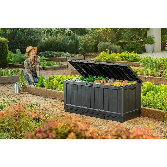 Keter Arcón para cojines de exterior Kentwood - L x An x Al: 128 x 53,6 x 59 cm, Plástico, Gris - Una caja de almacenamiento de jardín negra grande con tapa abierta, llena de herramientas de jardinería, está en el jardín.