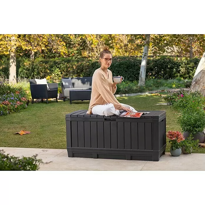 Keter Arcón para cojines de exterior Kentwood - L x An x Al: 128 x 53,6 x 59 cm, Plástico, Gris - Baúl de almacenamiento exterior gris oscuro texturizado, utilizado como banco en un patio, con una mujer sentada leyendo una revista.