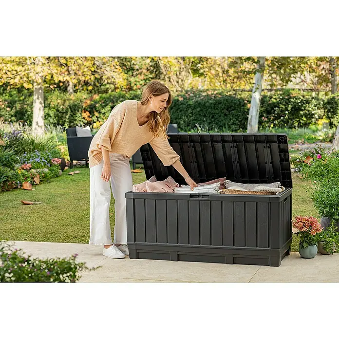 Keter Garten-Aufbewahrungsbox Kentwood - L x B x H: 128 x 53,6 x 59 cm, Kunststoff - Frau beugt sich zu einer schwarzen Gartenbox, um Decken zu holen. Praktische Aufbewahrungslösung für den Außenbereich.