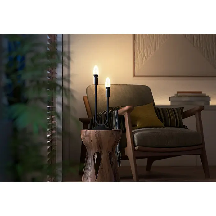 Philips Hue LED-Lampe Filament Classic White Ambiance - E14, 350 lm, Einstellbare Farbtemperatur, 1 Stk. - Nahaufnahme einer Hue Filament Kerze mit warmweissem Licht, die auf einem modernen Lampenfuss in einem gemütlichen Wohnraum steht.