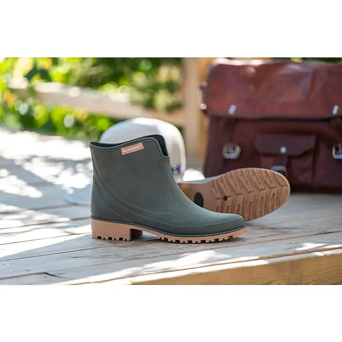 Black Fox Stiefelette Preston - Schuhgröße: 41, Khaki - Ein Paar Blackfox-Gummistiefel in Olivgrün mit braunen Sohlen, auf einer Holzoberfläche präsentiert und von einer Tasche und einem Hut begleitet.