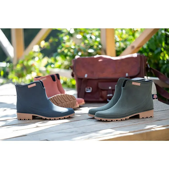 Black Fox Stiefelette PrestonSchuhgröße: 42, Blau - Drei Paare Blackfox-Gummistiefel in Marineblau, Rosa und Olivgrün auf einer Holzfläche, mit einer Ledertasche im Hintergrund.